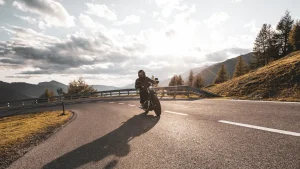 A person rides a motorcycle around a curved mountain road, with the sun low in the sky and trees and hills in the background.