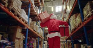 Person in a Santa Claus costume carries two gift boxes while walking through a warehouse aisle lined with stacked pallets and boxes.