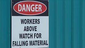 A sign reads "Danger: Workers above. Watch for falling material" on a teal metal wall.