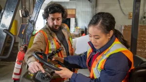 Two workers in safety vests inspect the controls of a forklift inside a warehouse. One points at the dashboard while the other looks closely at the steering wheel.