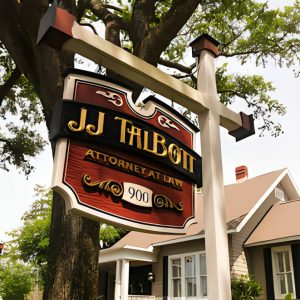 A wooden sign reading "JJ Talbott Attorney at Law 900" hangs from a post in front of a house with trees in the background.