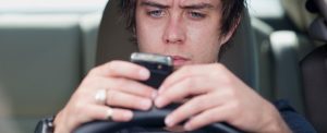 A man sits behind the wheel of a car, looking intently at his smartphone while holding it with both hands.