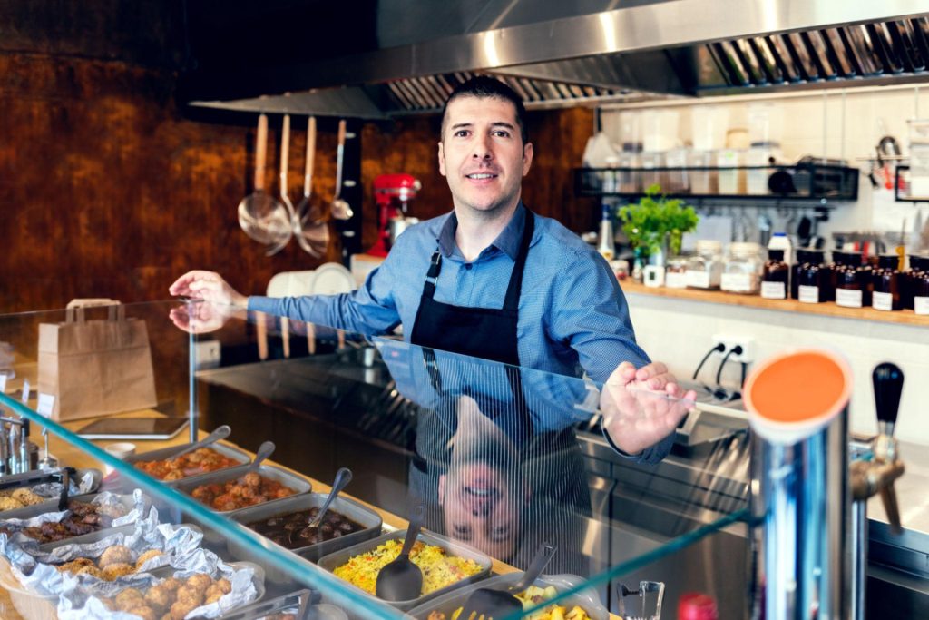 A man wearing a dark apron stands behind a glass counter displaying various prepared dishes in a restaurant kitchen.