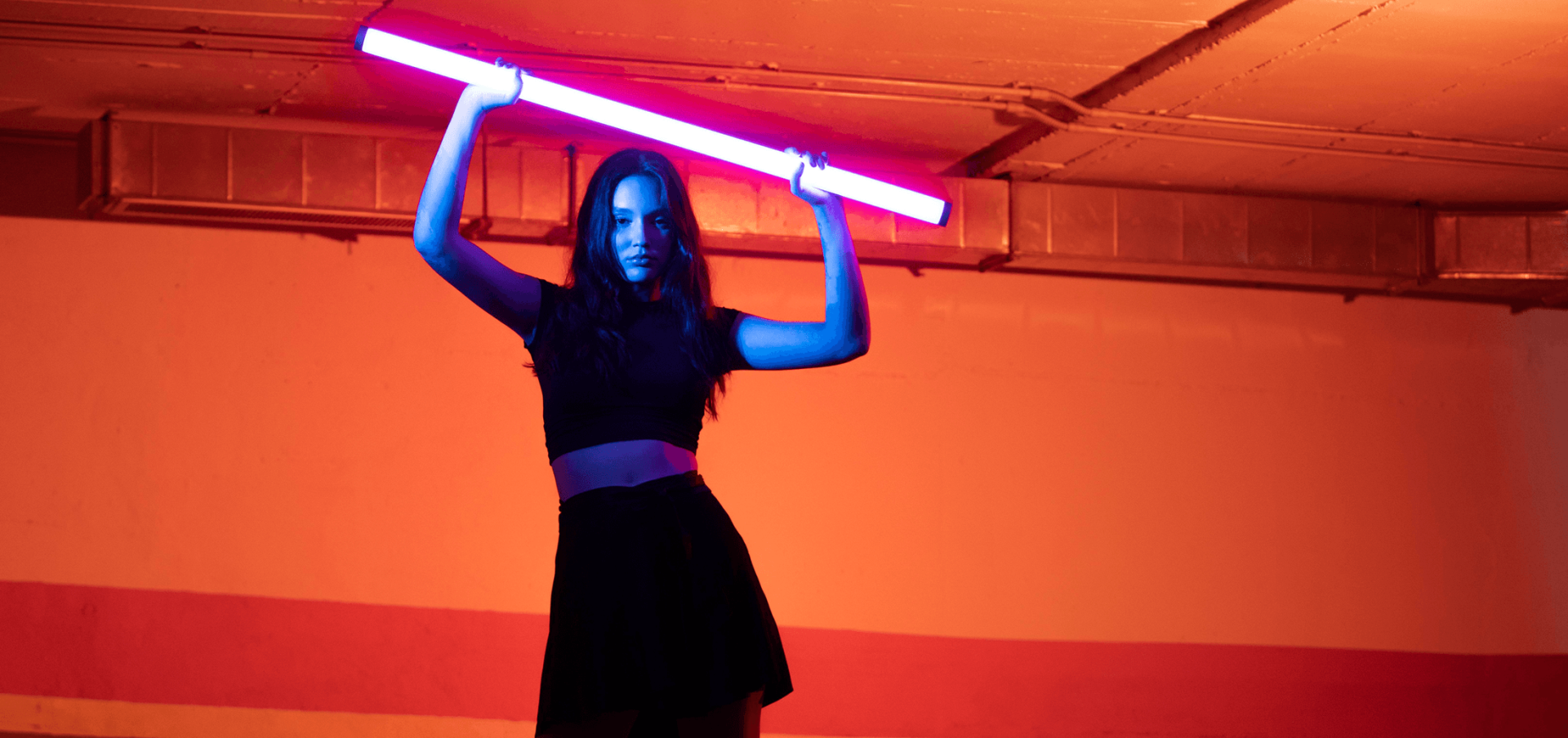 A woman in a black outfit stands in a brightly lit room, holding a glowing purple fluorescent light tube above her head.
