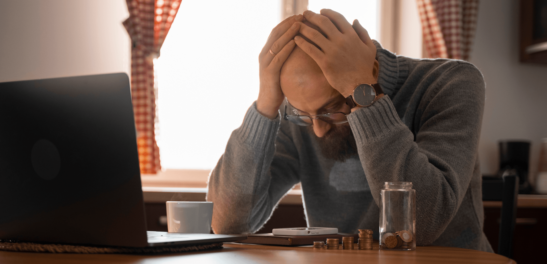 A man sits at a table with his head in his hands, looking stressed. Coins, a jar, a mug, and a laptop are on the table in front of him.