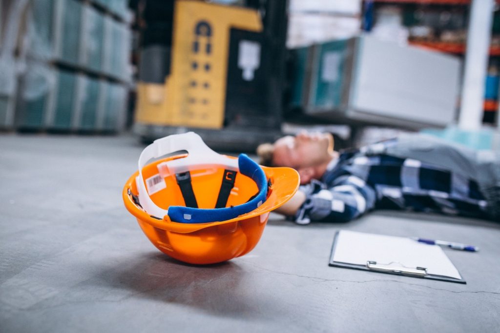 An orange safety helmet lies on the ground in a warehouse, with a person in the background lying on the floor near a clipboard and pen.