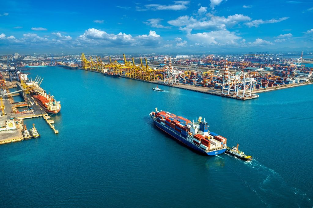 A large cargo ship travels through a busy port with multiple cranes, containers, and ships under a bright blue sky.