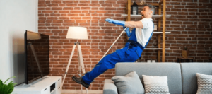 A man in blue overalls slips backward over a rope in a living room, near a couch, TV, and floor lamp, with a surprised expression on his face.