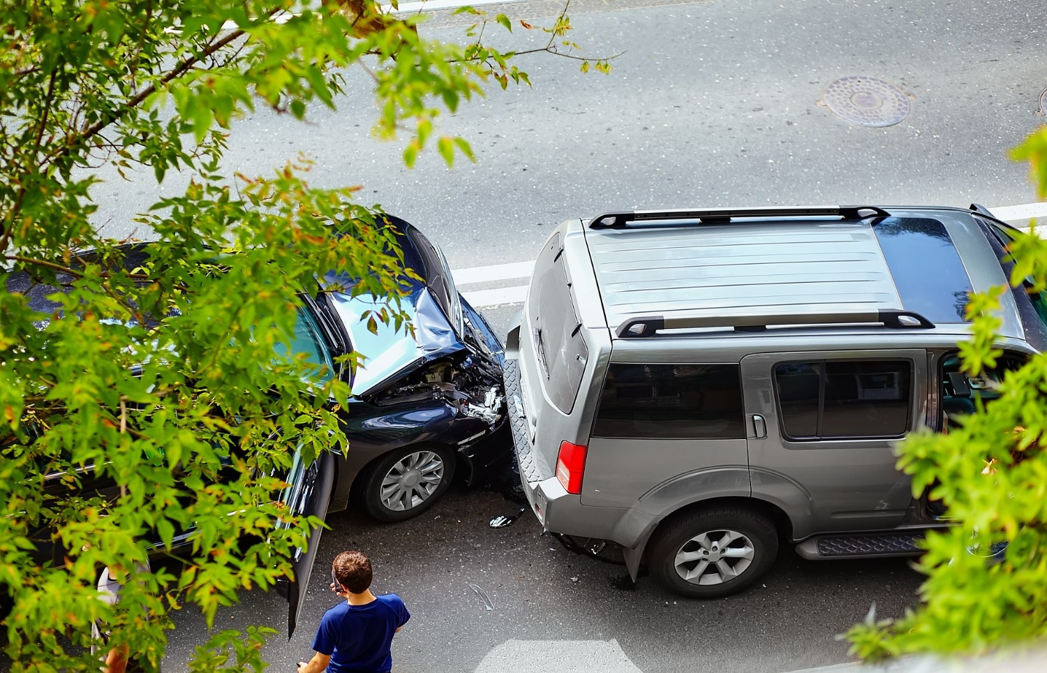 A black car has rear-ended a gray SUV on a city street. A person stands nearby, and green leafy branches partially frame the scene.