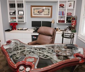 A modern office with a brown leather chair, a large decorative glass-and-metal desk, dual monitors, shelves with books and memorabilia, and a window with blinds.