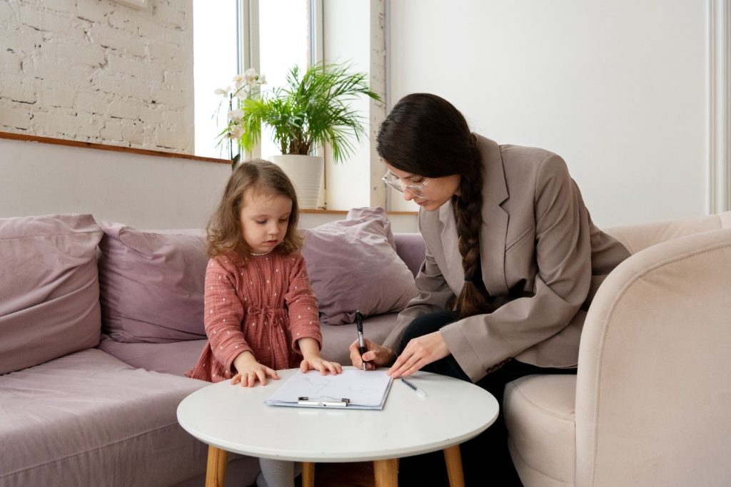A woman in a blazer sits on a couch next to a young girl, pointing at a clipboard with papers on a coffee table in front of them.