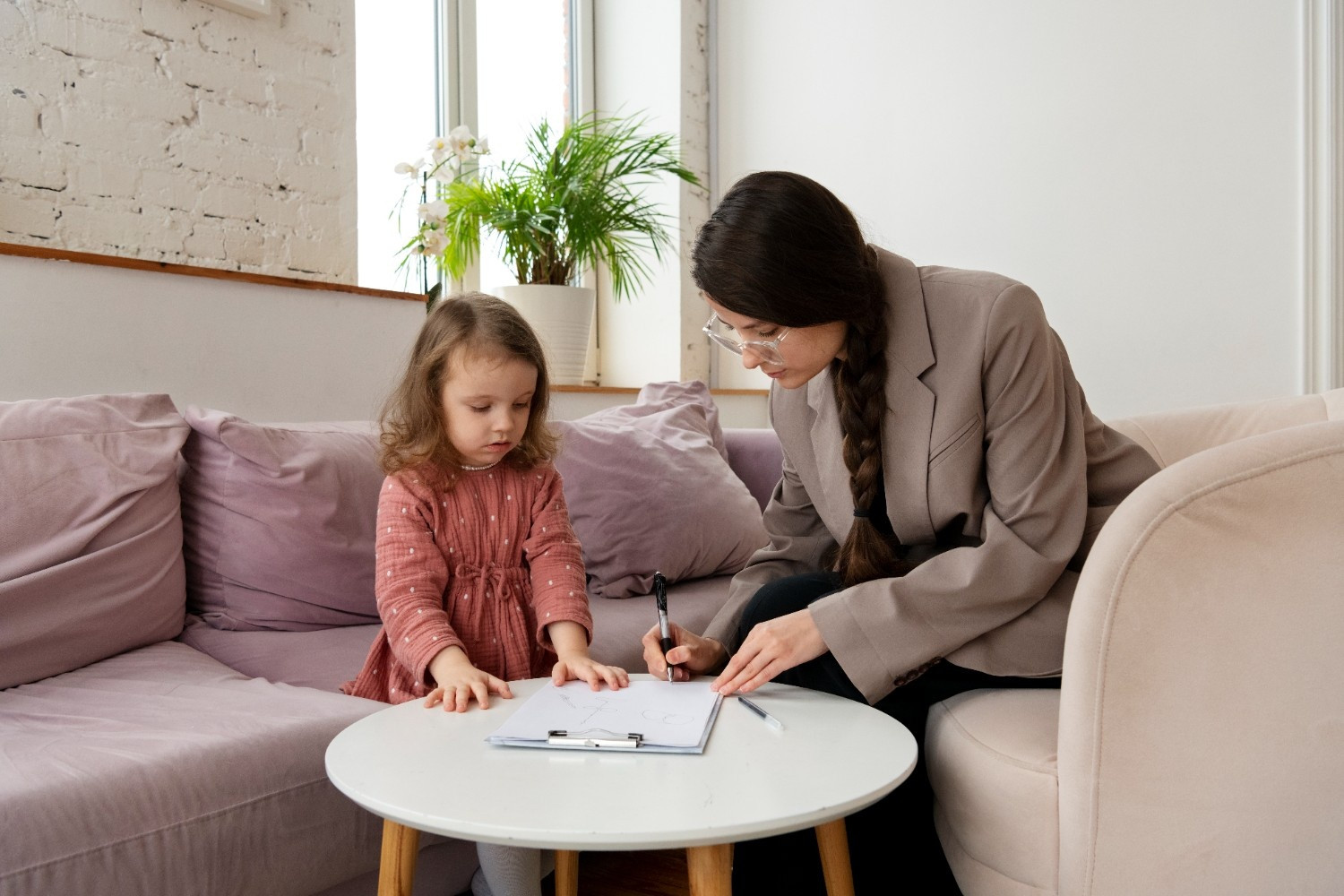 A woman in a blazer sits on a couch next to a young girl, pointing at a clipboard with papers on a coffee table in front of them.