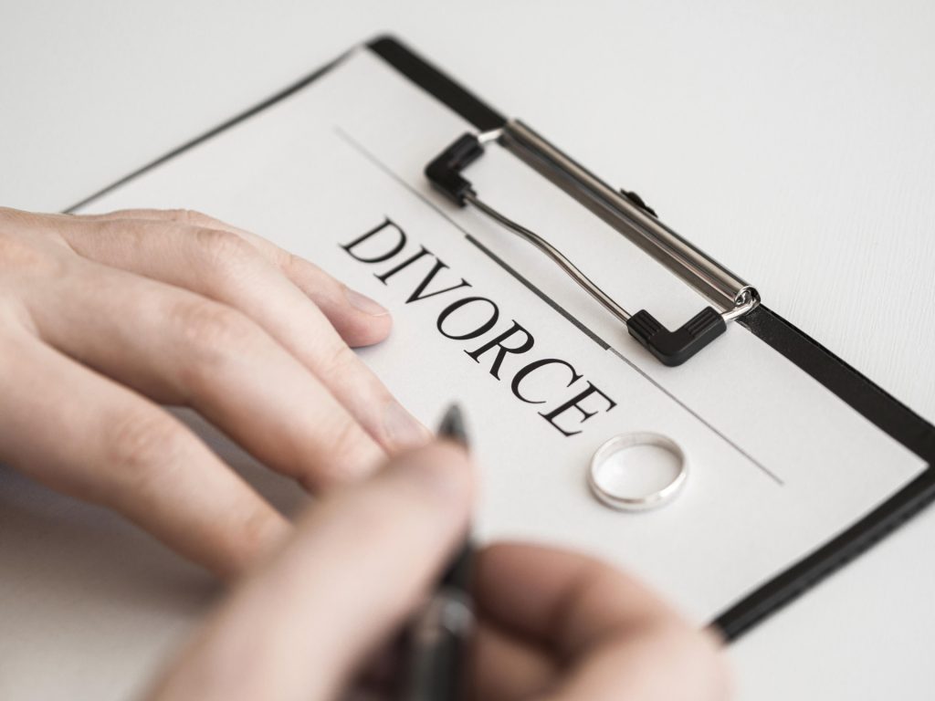 A person prepares to sign a divorce document on a clipboard, with a wedding ring placed on the paper.