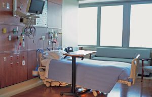 Empty hospital room with a neatly made bed, medical equipment on the wall, a monitor, large windows, and a sofa in the background.