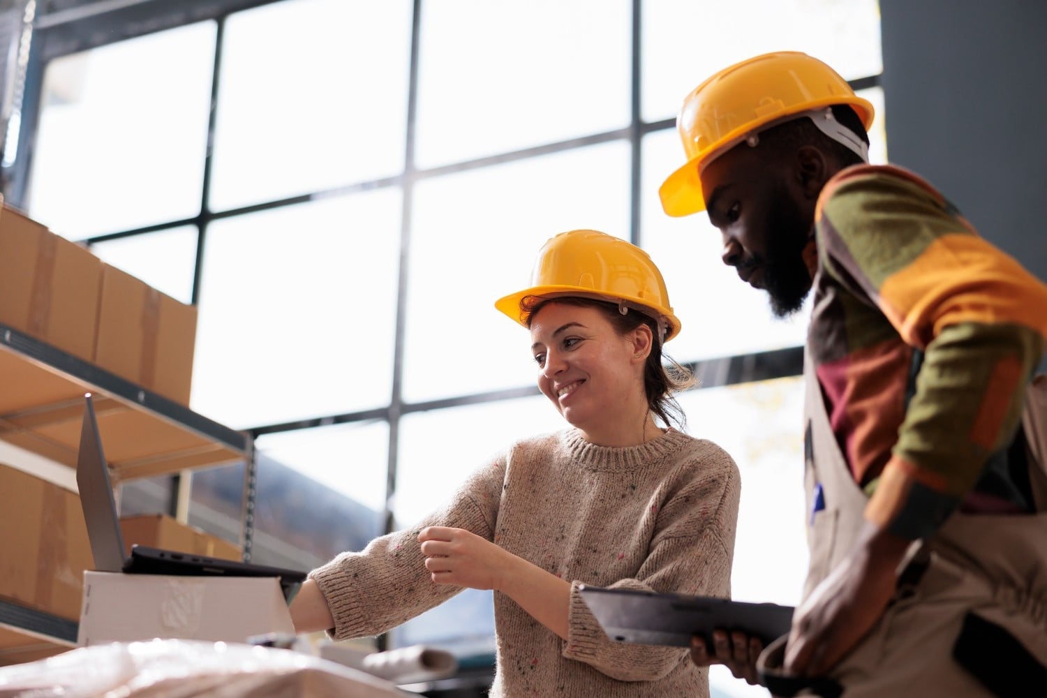 Two workers wearing yellow hard hats look at a laptop in a warehouse, with shelves and large windows visible in the background.