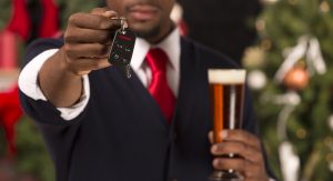 A man in a suit holds a car key in one hand and a glass of beer in the other, with a decorated Christmas tree in the background.