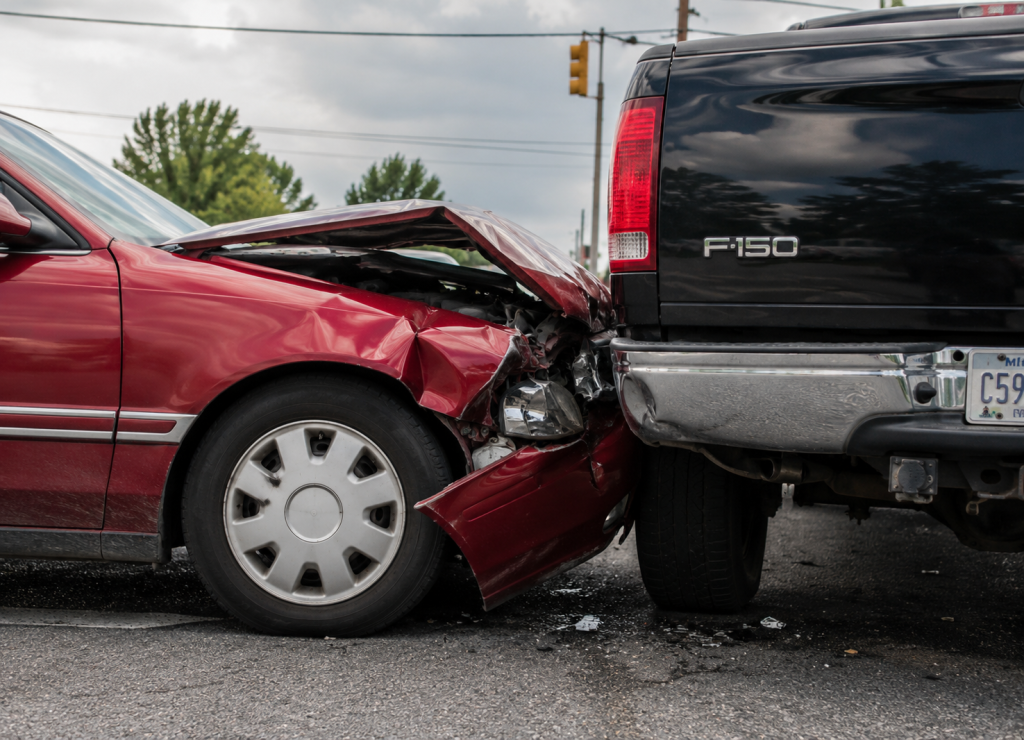 A red sedan with a damaged front end after rear-ending a black Ford F-150 pickup truck at an intersection.