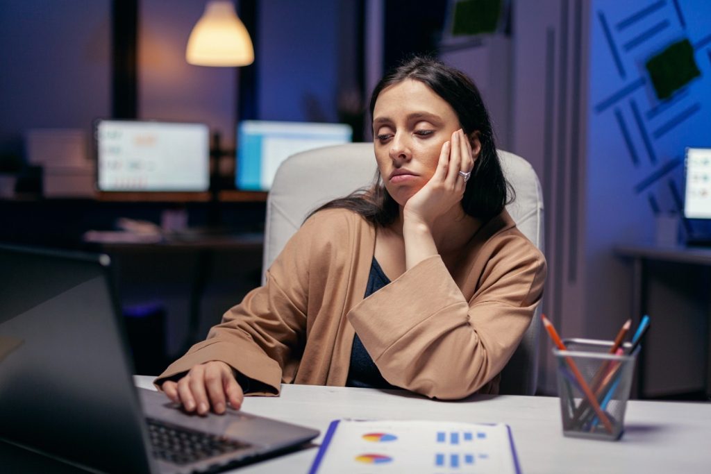 A woman sits at a desk, looking tired and bored while staring at a laptop screen in an office setting at night.
