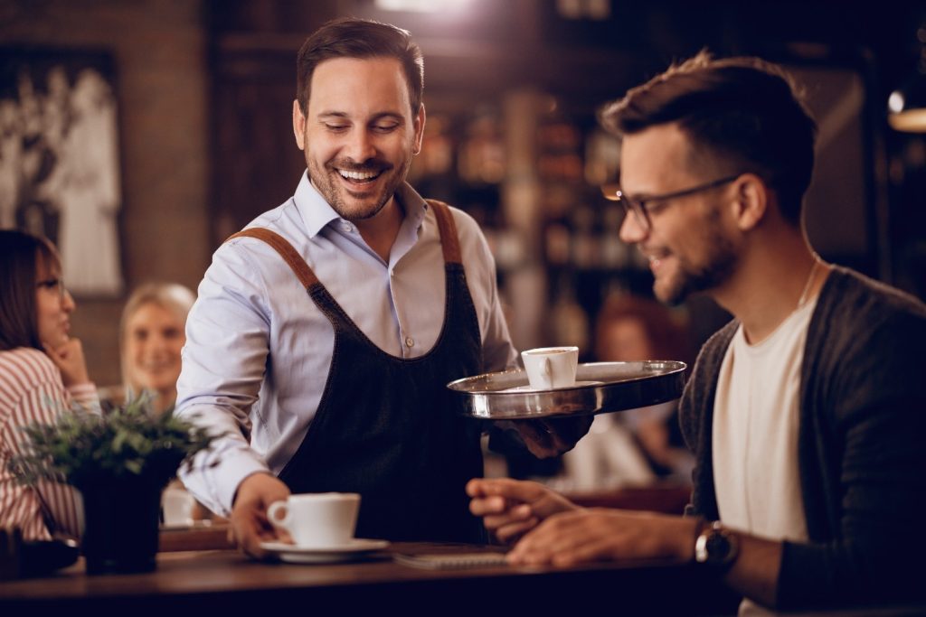 A smiling waiter serves coffee to a seated man at a café, with other customers visible in the blurred background.