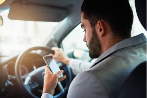 A man sits in the driver's seat of a car, holding the steering wheel with one hand and using a smartphone with the other hand.