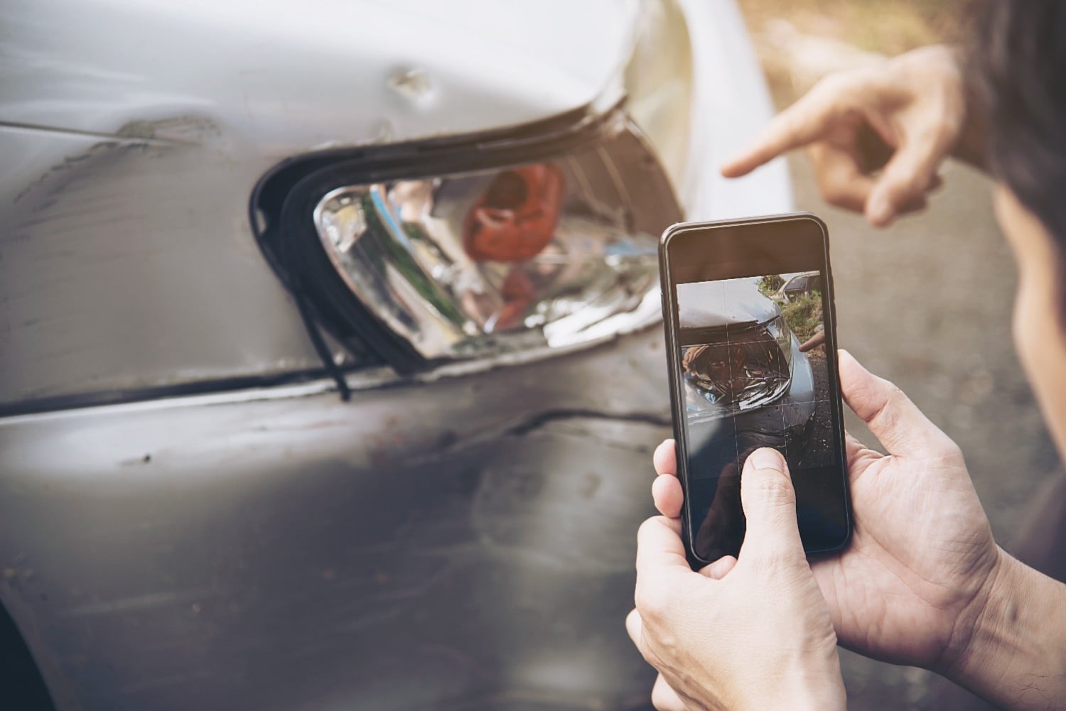 A person takes a photo of damage to a car's front bumper and headlight with a smartphone while another person points at the damage.