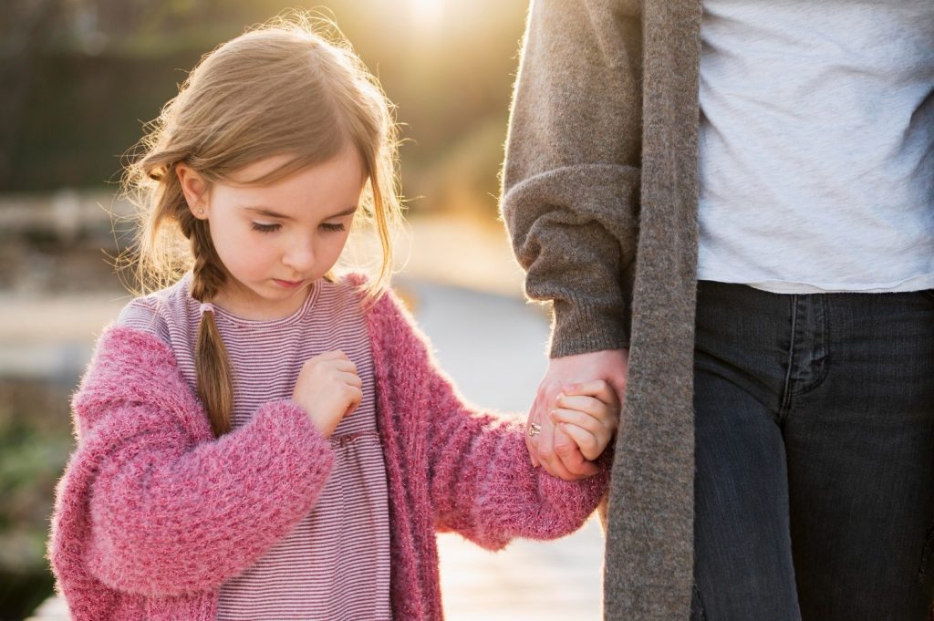 A young girl in a pink sweater holds hands with an adult wearing a gray cardigan, standing outdoors in sunlight.