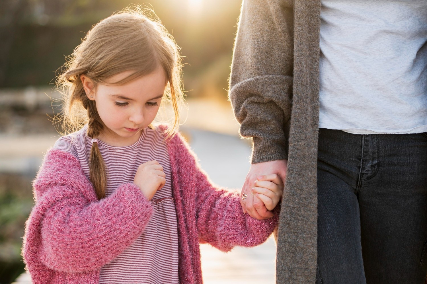 A young girl in a pink sweater holds hands with an adult wearing a gray cardigan, standing outdoors in sunlight.