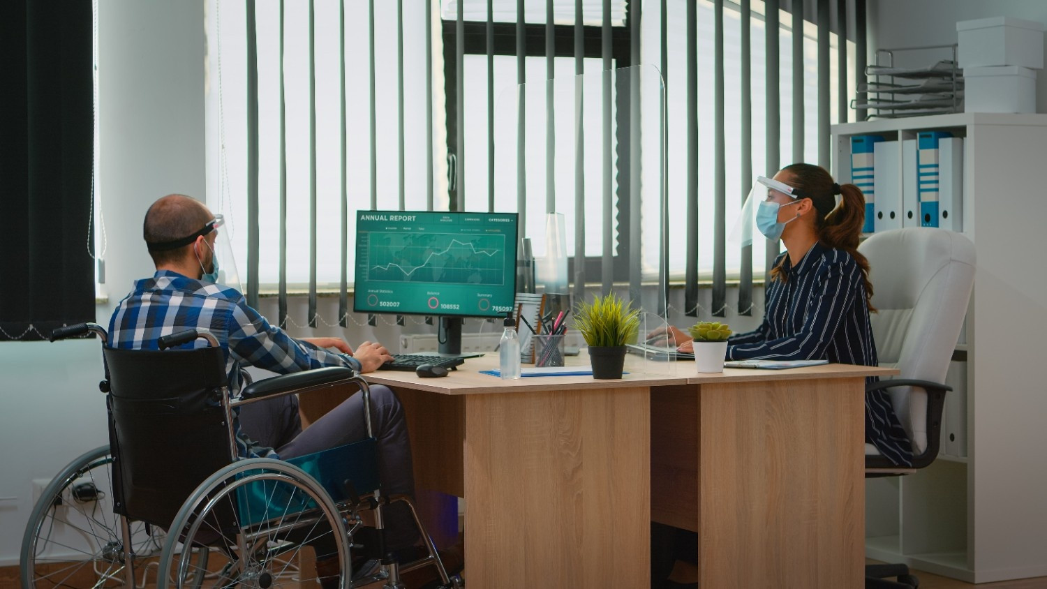 Two people wearing face masks and shields sit across from each other at an office desk; one uses a wheelchair, and a computer screen shows an annual report with charts.