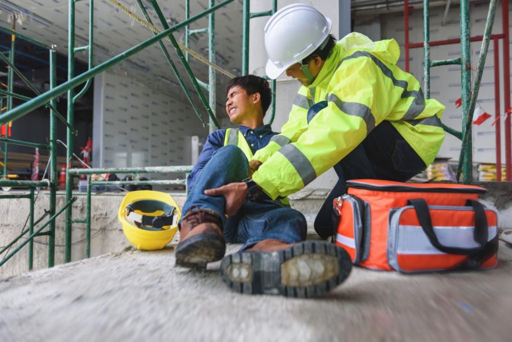 A construction worker sits on the ground holding his leg in pain while another worker in safety gear assists him; a first aid kit and helmet are nearby.