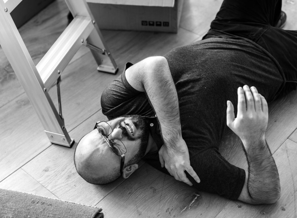 A man lies on the floor near a ladder, clutching his shoulder and appearing to be in pain, suggesting a possible fall or injury at home.