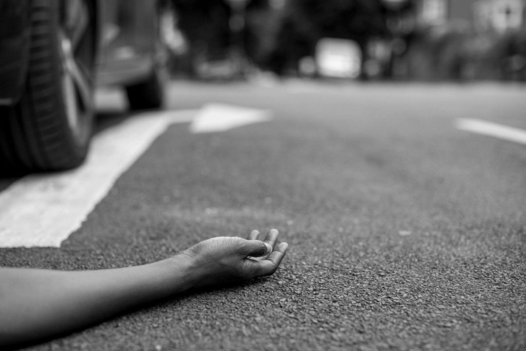 A person's arm lies on the pavement near a car, with the hand extended and palm facing up; the scene appears to suggest an accident.
