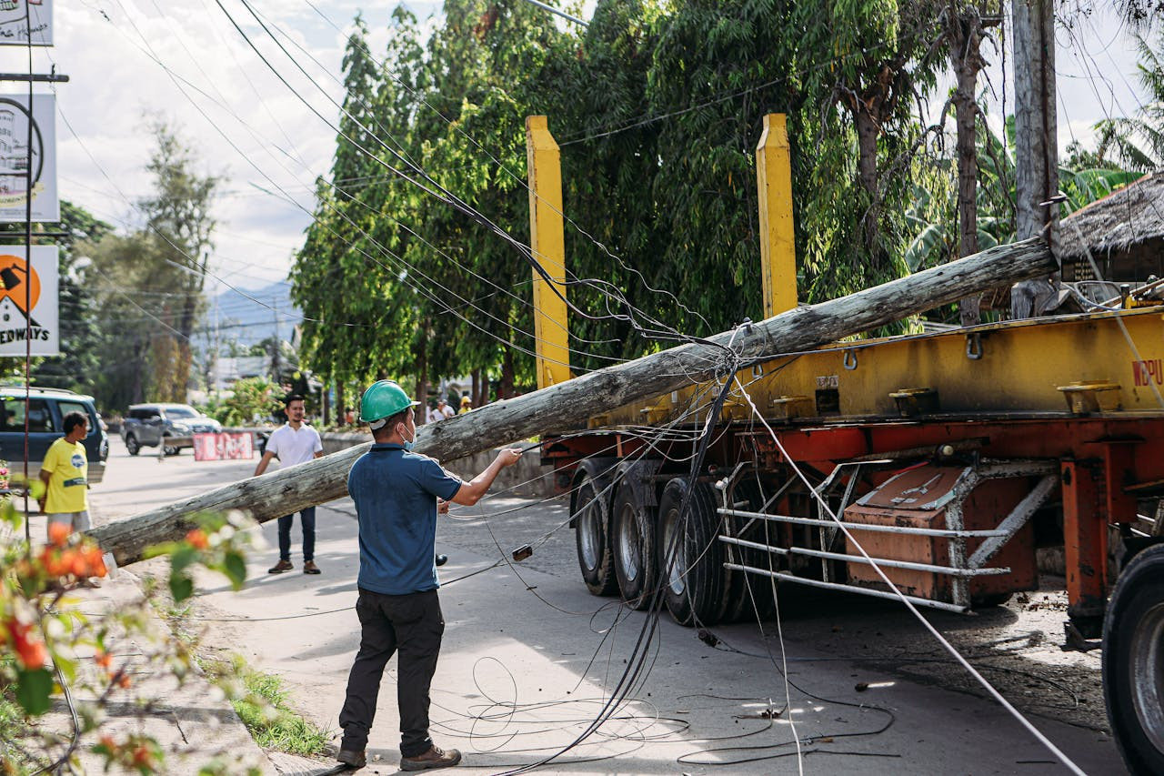 A worker in a green helmet steadies a fallen utility pole tangled in wires as it rests on a truck trailer on a city street.
