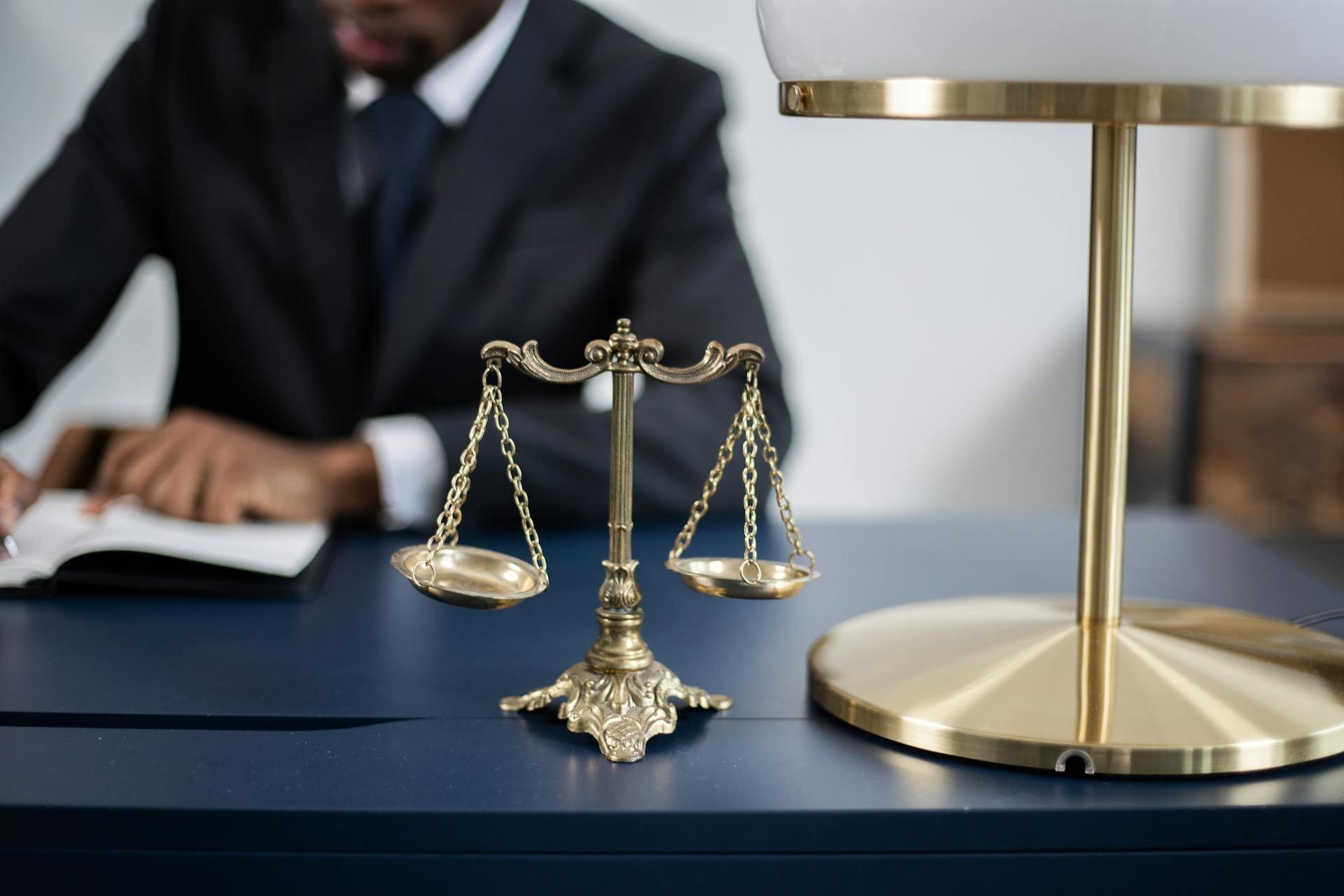 A brass balance scale and lamp sit on a blue desk, with a person in a suit writing in the background.
