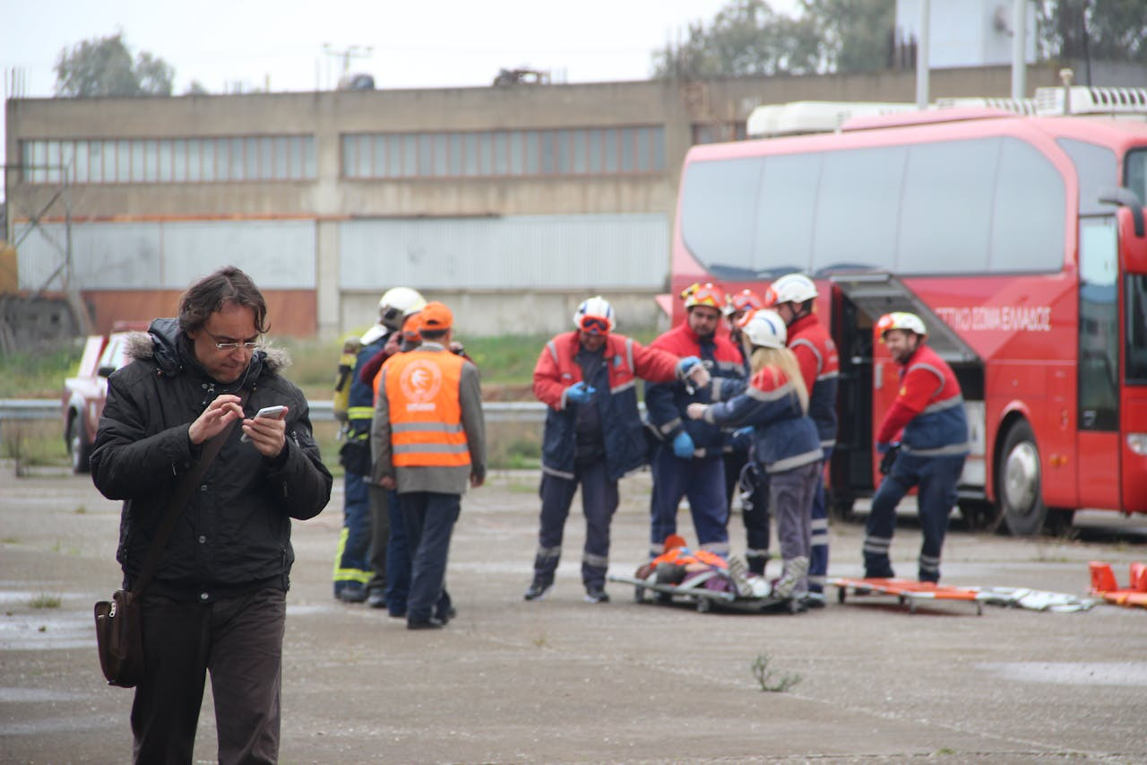 A man walks in the foreground checking his phone as emergency responders assist someone near a stretcher beside a red bus in an industrial area.