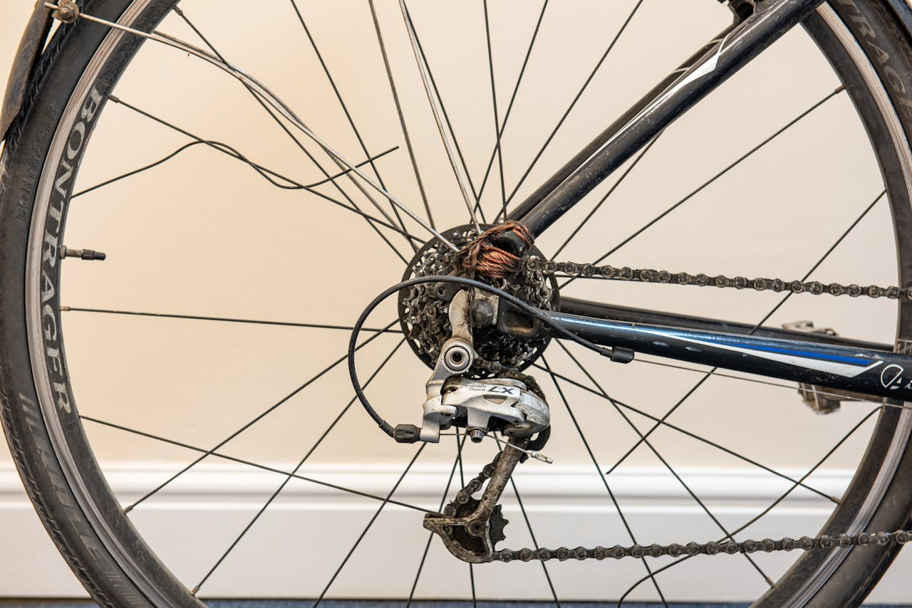 Close-up of a bicycle’s rear wheel, showing the gear cassette, derailleur, and part of the black frame against a beige wall.