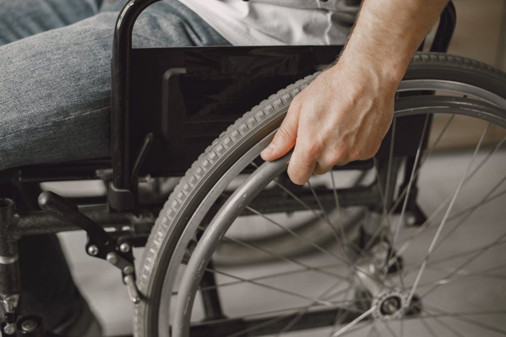 Close-up of a person’s hand gripping the wheel of a manual wheelchair, with part of their torso and jeans visible.