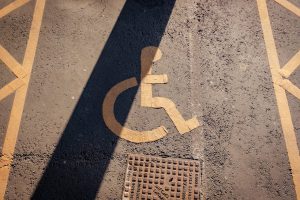 Yellow wheelchair-accessible symbol painted on asphalt, partially covered by a dark shadow, with yellow diagonal parking lines and a drainage grate visible.