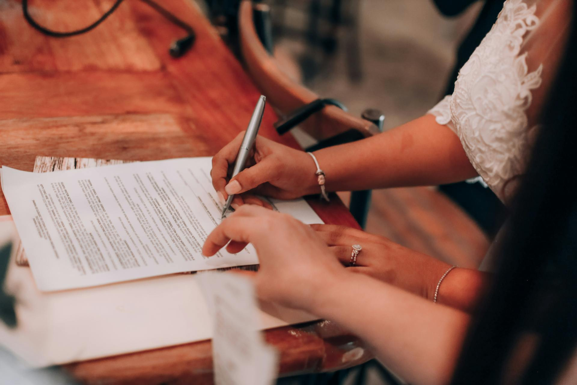 Two people sign a printed document on a wooden table; one person holds the paper while the other writes with a pen.