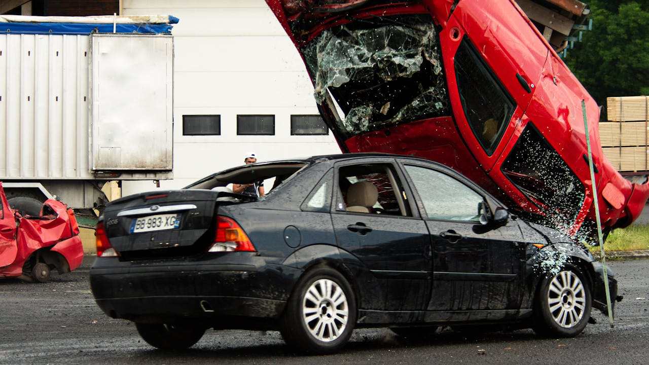 A black car with a damaged rear and broken windows is parked near a red car that is overturned and heavily damaged, with shattered glass scattered around.