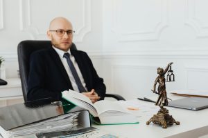 A man in a suit sits at a desk with legal documents, an open book, newspapers, and a Lady Justice statue in an office setting.
