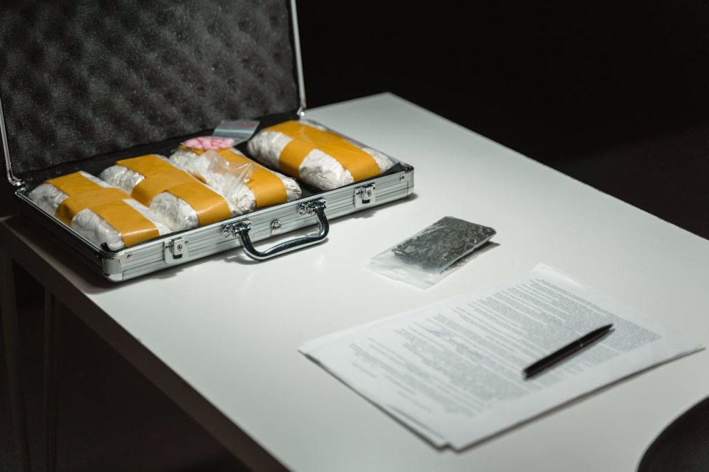 A metal briefcase containing wrapped packages, a plastic bag with a dark substance, and documents with a pen are placed on a white table.