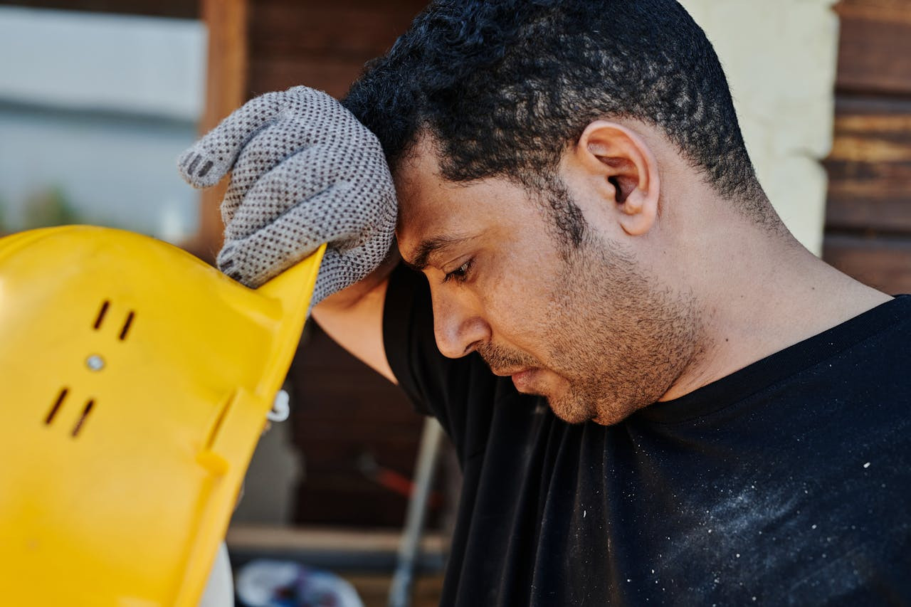 A construction worker wearing a glove holds a yellow hard hat to his forehead, appearing tired or overheated.