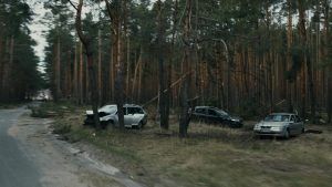 Three damaged cars are abandoned on the side of a road in a forested area, with fallen branches and debris scattered around.