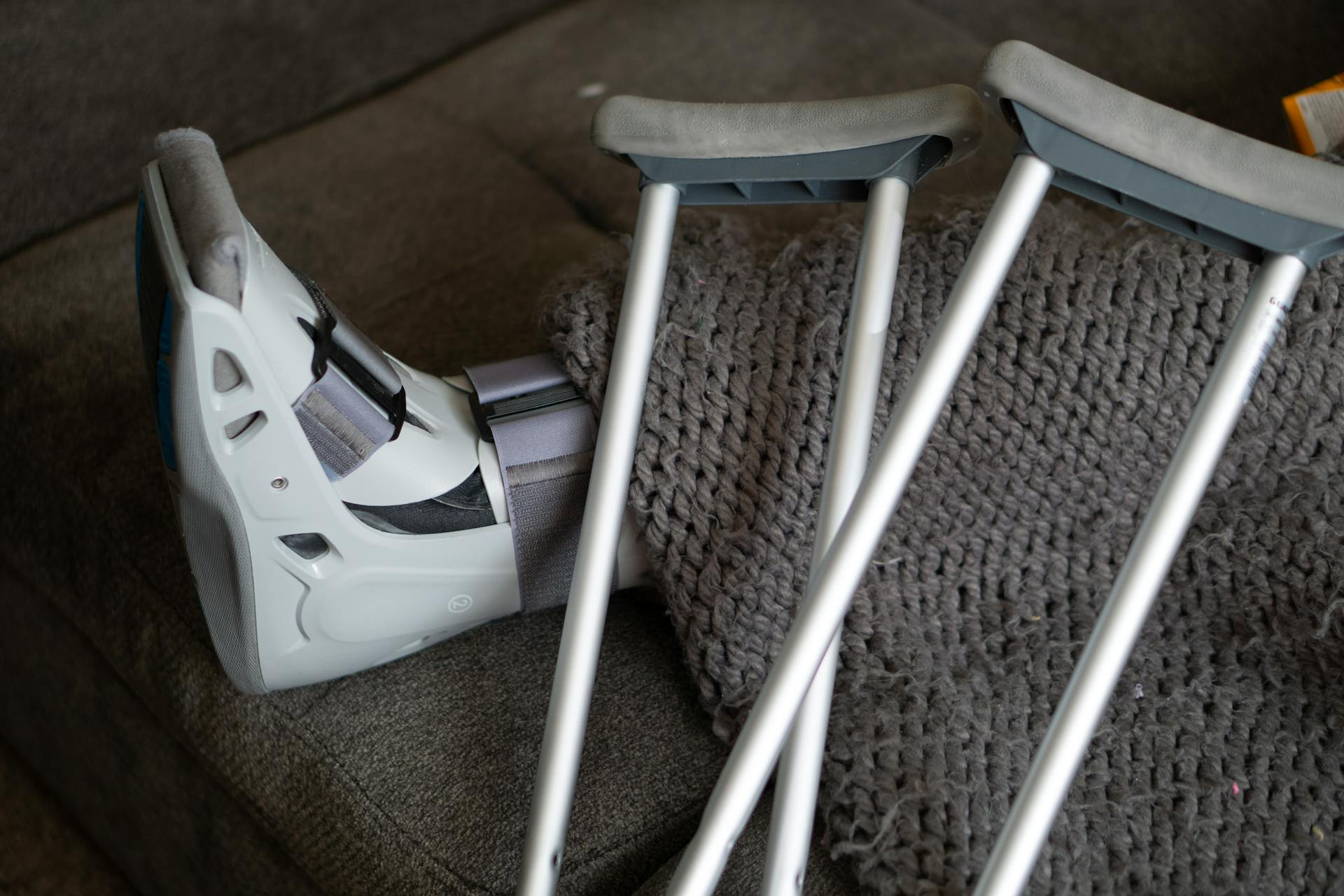 A person with a medical boot on their foot rests on a couch with two crutches and a knitted gray blanket beside them.