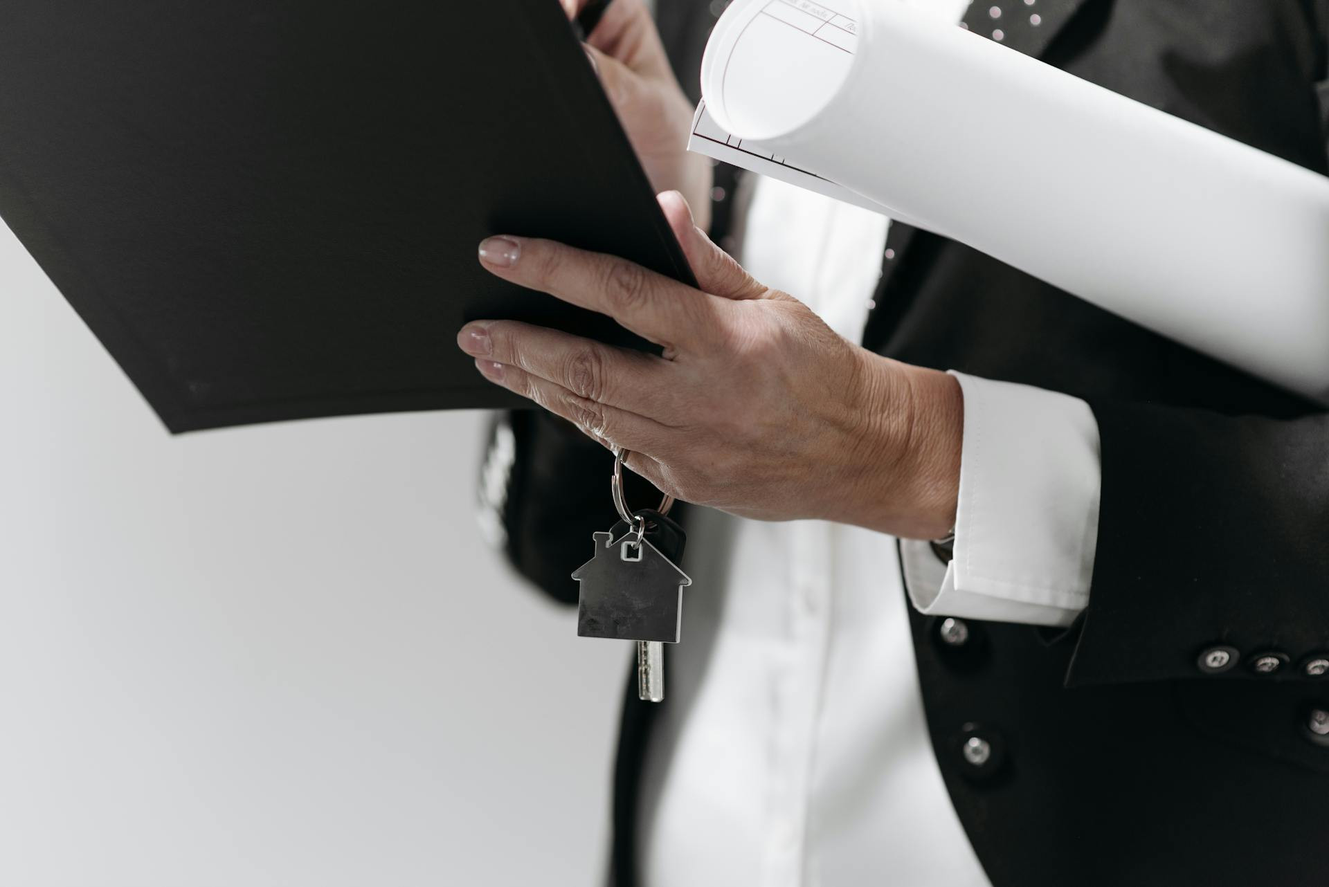 Person in a suit holding a clipboard, rolled documents, and house-shaped keys, suggesting activities related to real estate or property transactions.