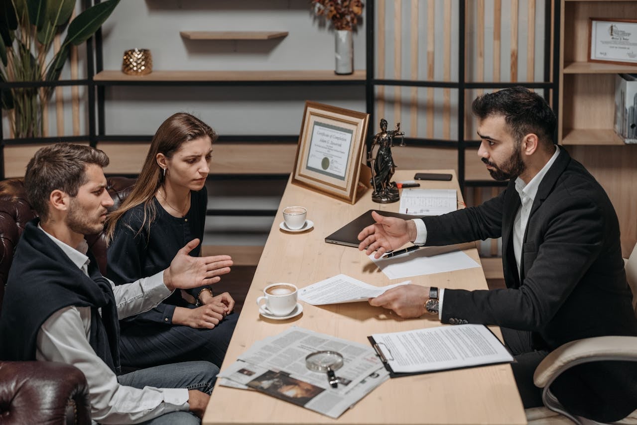 A man and woman sit across a desk from a professional holding papers, engaged in discussion in an office setting with documents and coffee cups on the table.