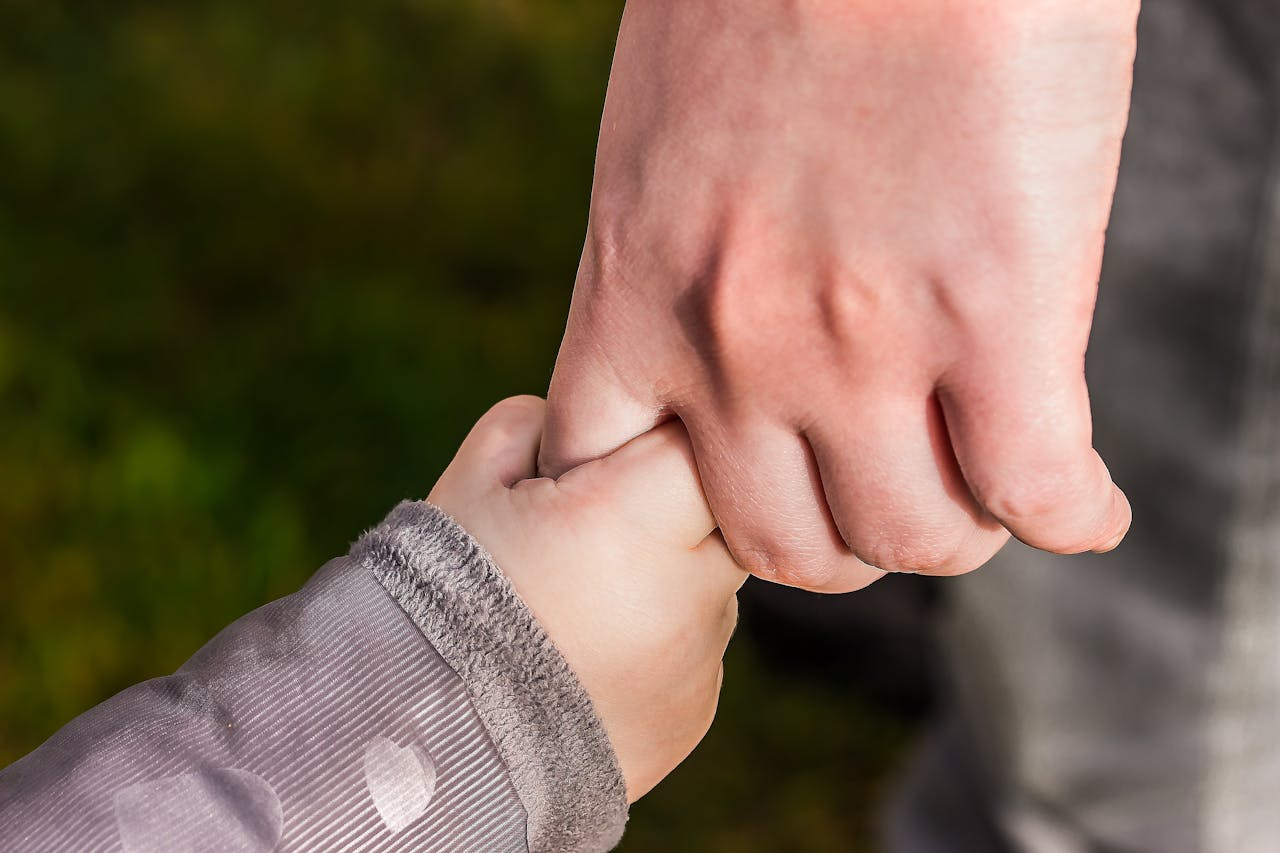 An adult hand gently holds a child's hand outdoors, with the child wearing a light grey jacket and the background blurred.