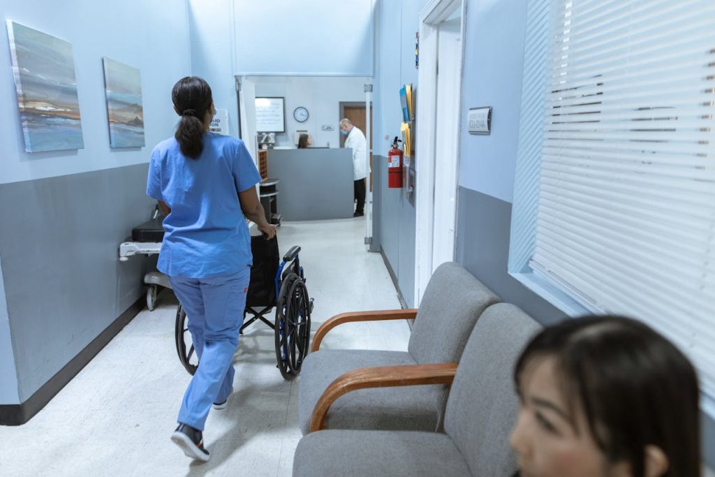 A healthcare worker in blue scrubs pushes an empty wheelchair down a hallway in a medical facility, while a woman sits in the waiting area.