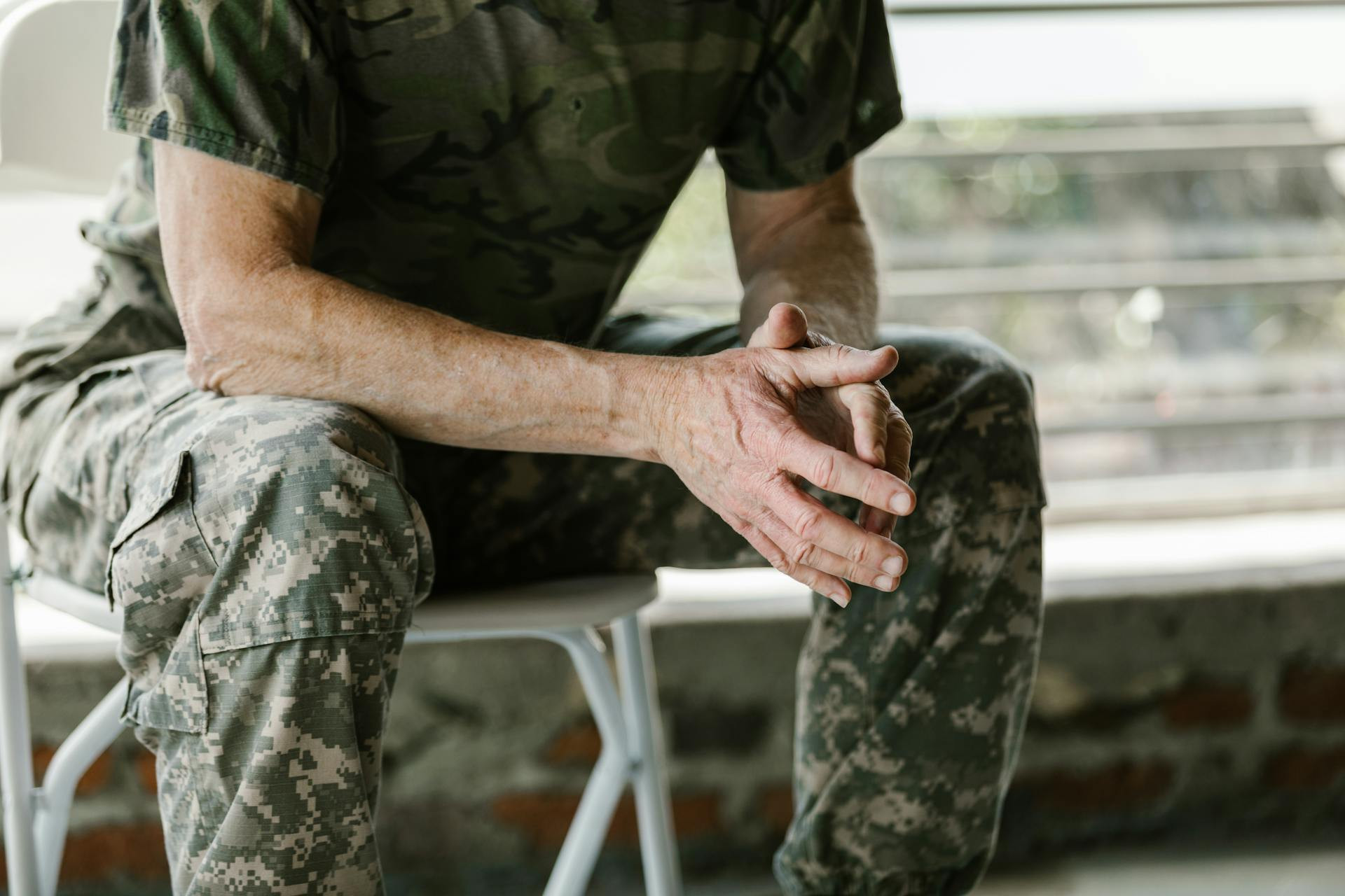 A person in camouflage military uniform sits on a white chair with their hands clasped, only their torso, arms, and legs visible.
