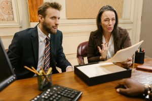 Two people in business attire sit at a desk reviewing documents, with one person gesturing and speaking, while another listens attentively.